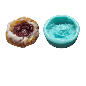 May include: A close-up of a pastry with a white glaze and a red filling. The pastry is next to a blue silicone mold.