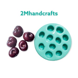 May include: A turquoise silicone mold with 12 round cavities, perfect for making small treats like chocolates or candies.  Five dark red cherries are scattered on a white surface next to the mold.