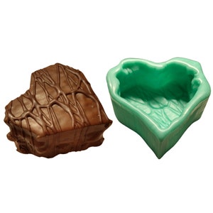 May include: A heart-shaped chocolate dessert with a dark chocolate drizzle next to a teal heart-shaped mold. The dessert has a rectangular shape with rounded edges. The mold is empty and has a textured interior.