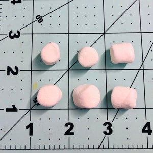 Pu&ograve; includere: Sei piccoli marshmallow cilindrici di colore rosa pallido disposti in due file su un tappetino da taglio azzurro con linee a griglia e numeri neri. I marshmallow sono posizionati contro un righello con misure in pollici, indicando una dimensione di 1,3 cm.