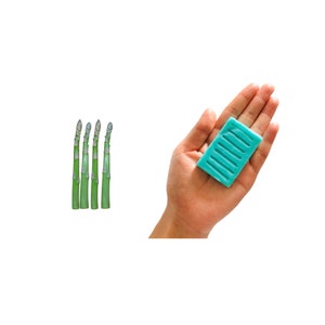 May include: Five green asparagus spears with purple tips stand upright on a white background. A hand holds a light blue rectangular bar of soap with ridges.