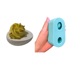 May include: A light blue silicone mold for making deviled eggs. The mold has two cavities, each shaped like a deviled egg. The mold is shown in a hand.