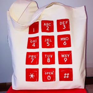 Op de afbeelding: Een crèmekleurige canvas tas met een grote, rode print van een telefoontoetsenbord. Het ontwerp bestaat uit twaalf vierkante knoppen in een raster, elk voorzien van witte cijfers, letters en symbolen, waaronder het sterretje en hekje.