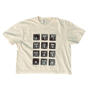 Op de afbeelding: Een crèmekleurig T-shirt met korte mouwen en een grafische print van een telefoontoetsenbord. Het ontwerp toont zwarte vierkante toetsen met witte cijfers, letters en symbolen in een raster, inclusief de standaard 0-9, sterretje en hekje.