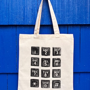Op de afbeelding: Een beige canvas draagtas met een zwarte grafische print van een standaard telefoontoetsenbord. Het ontwerp toont de cijfers één tot en met negen, nul, het sterretje, het hekje en de bijbehorende letters gerangschikt in een raster.