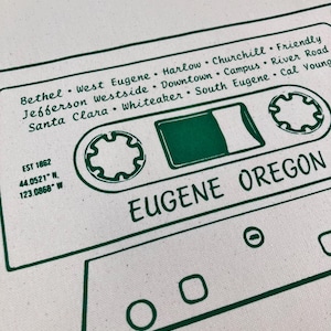 Op de afbeelding: Witte draagtas met een groen cassettebandontwerp. Het ontwerp bevat de tekst "Eugene Oregon" en namen van buurten. De draagtas heeft ook de tekst "EST 1862" en coördinaten 44.0521° N, 123.0868° W.