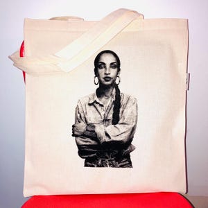 Op de afbeelding: Een beige canvas draagtas met een zwart-witte grafische print van een persoon met een lange vlecht, grote oorringen en een overhemd met gekruiste armen. De tas heeft twee bijpassende beige stoffen hengsels en een klein label aan de zijkant.