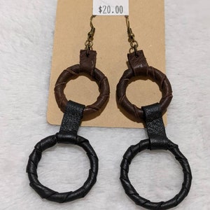 May include: A pair of handmade earrings with a brown and black leather design. The earrings feature a circular shape with a braided texture. The top portion is brown, and the bottom is black. The earrings hang from bronze-colored hooks.