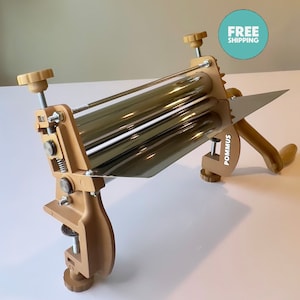 May include: A tan pasta maker with a silver roller and a hand crank. The machine has a clamp to secure it to a table. The brand name "POMMUS" is printed on the side. The image also includes a "FREE SHIPPING" badge.