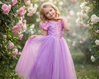 May include: A young girl in a lavender princess dress with a bodice and a flowing tulle skirt. The dress has pink accents and short sleeves. She is standing in a garden surrounded by pink and white roses.