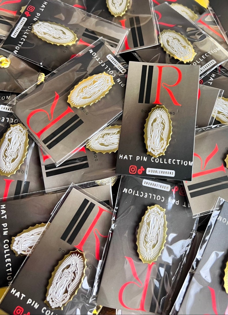 Może przedstawiać: Zbliżenie na stos kart z przypinką ze złota i białego emaliowanego przedstawiającą postać religijną. Na kartach widnieje napis "HAT PIN COLLECTION" i "DOBLERBRAND".