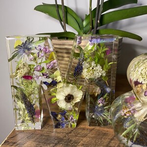 May include: Two clear resin letters, "M" and "W", filled with pressed flowers and greenery. The flowers are in shades of white, purple, pink, and green. The letters are sitting on a wooden surface.