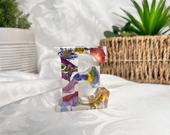 May include: A clear resin letter E with pressed flowers and petals inside. The letter is a light pink, blue, yellow, and green colour.
