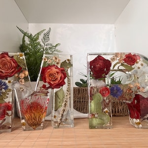 May include: Two clear resin letters, "M" and "R", filled with pressed flowers and greenery. The flowers are mostly red and pink roses, with some white and blue flowers.
