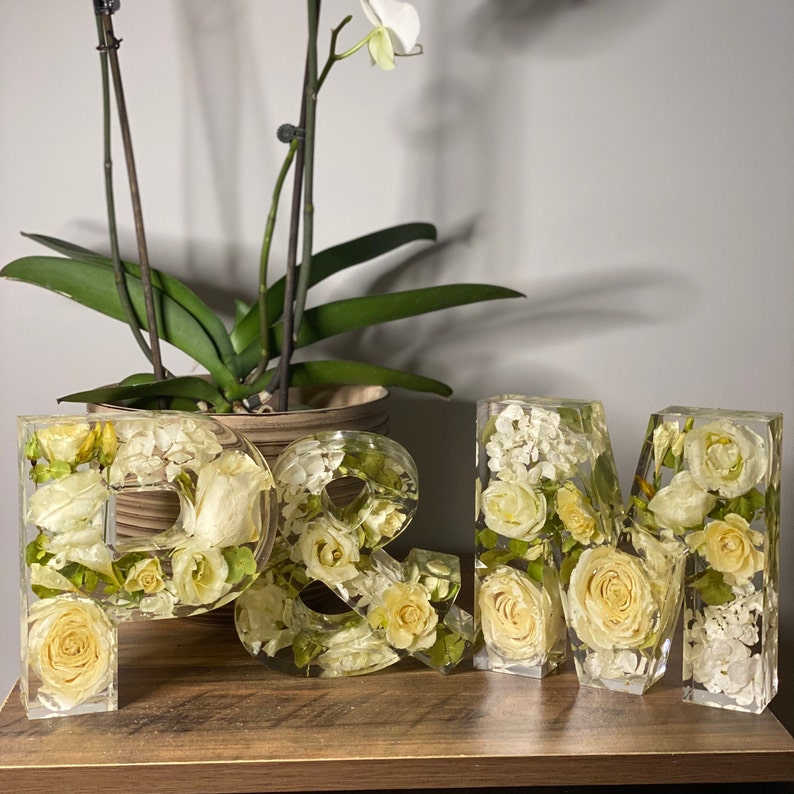 May include: Three white resin letters, "P", "&", and "M", filled with pressed white flowers and greenery. The letters are arranged in a row on a wooden surface.