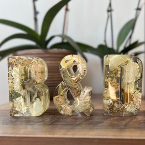 May include: Three clear resin letters, "B", "&", and "D", filled with dried white and cream flowers and greenery. The letters are sitting on a wooden surface.