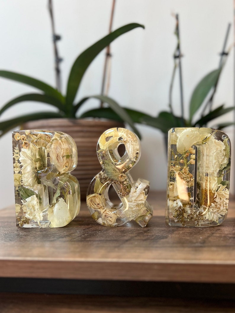 May include: Three clear resin letters, "B", "&", and "D", filled with dried white and cream flowers and greenery. The letters are sitting on a wooden surface.