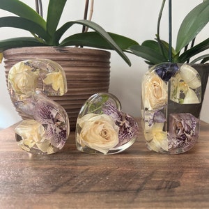 May include: Three resin ornaments shaped like the letters 'S', 'G', and a heart, each filled with dried flowers and leaves. The flowers are white and purple, and the leaves are green.
