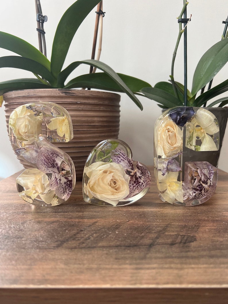 May include: Three resin ornaments shaped like the letters 'S', 'G', and a heart, each filled with dried flowers and leaves. The flowers are white and purple, and the leaves are green.