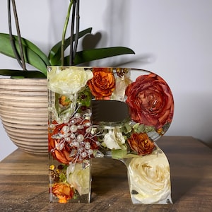 May include: A clear resin letter "R" filled with dried flowers in shades of white, orange, and red. The flowers are arranged in a random pattern and include small white beads.
