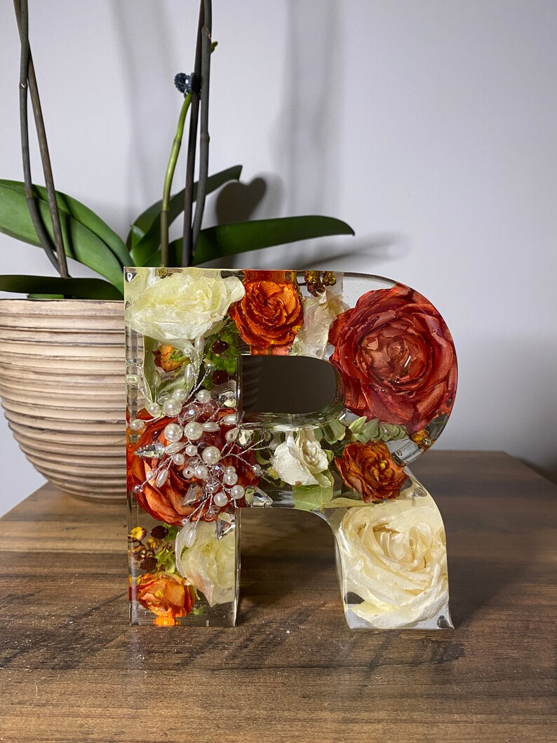 May include: A clear resin letter "R" filled with dried flowers in shades of white, orange, and red. The flowers are arranged in a random pattern and include small white beads.