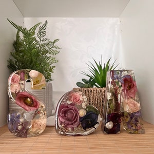 May include: Three resin letters spelling "S A A" with dried flowers and greenery encased in the resin. The letters are on a wooden surface.