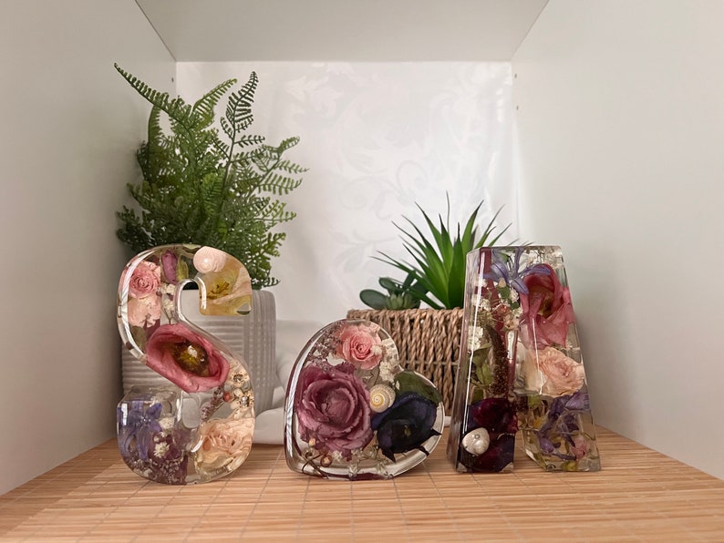 May include: Three resin letters spelling "S A A" with dried flowers and greenery encased in the resin. The letters are on a wooden surface.