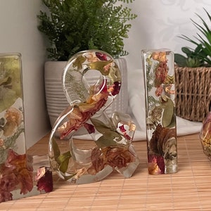 May include: A set of four clear resin letters, spelling out "& I", with dried flowers and leaves embedded inside. The letters are standing on a light brown surface.