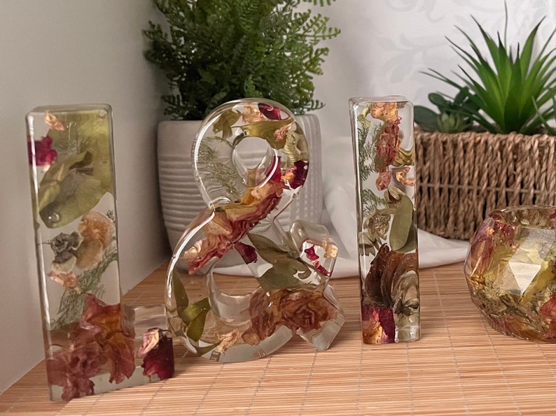 May include: A set of four clear resin letters, spelling out "& I", with dried flowers and leaves embedded inside. The letters are standing on a light brown surface.