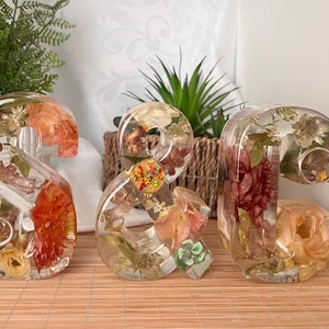 May include: Three resin numbers, 2, & and 2, filled with dried flowers and leaves in shades of orange, yellow, green, and brown. The numbers are on a light brown wood surface.