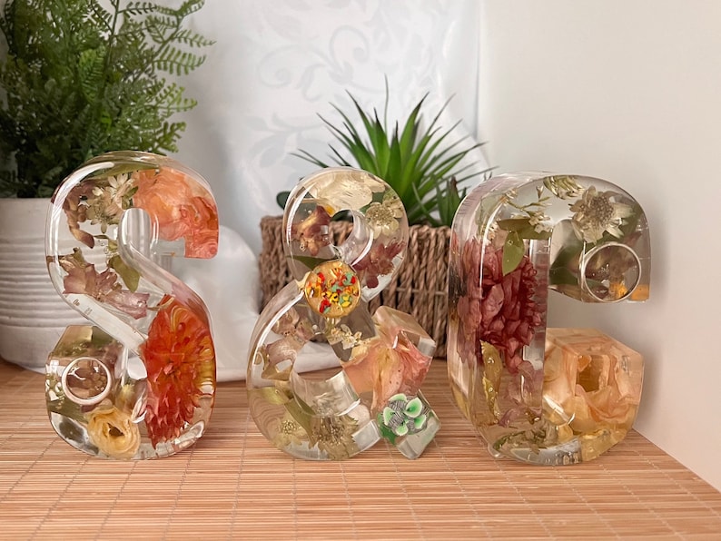May include: Three resin numbers, 2, & and 2, filled with dried flowers and leaves in shades of orange, yellow, green, and brown. The numbers are on a light brown wood surface.