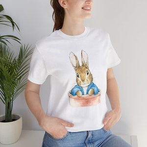 Può includere: T-shirt bianco con un'illustrazione ad acquerello di un coniglio marrone con una giacca blu, seduto in un vaso di terracotta. Il coniglio ha lunghe orecchie e un viso dettagliato. La maglietta è a girocollo classico.