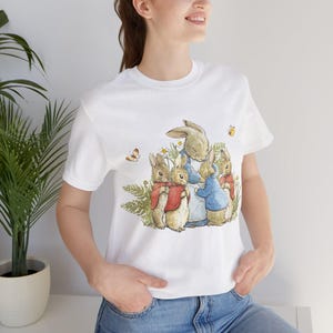 Puede incluir: Camiseta blanca con un estampado de Peter Rabbit y su familia. Los conejos están rodeados de flores y mariposas.