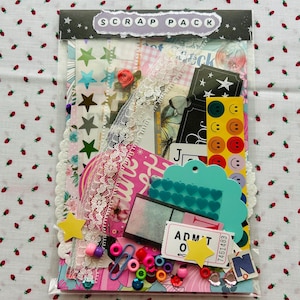 May include: A clear plastic bag filled with scrapbooking supplies. The bag contains various embellishments, including star stickers, lace trim, colorful beads, paper clips, and die-cut shapes. The top of the bag has a label that reads "SCRAP PACK".