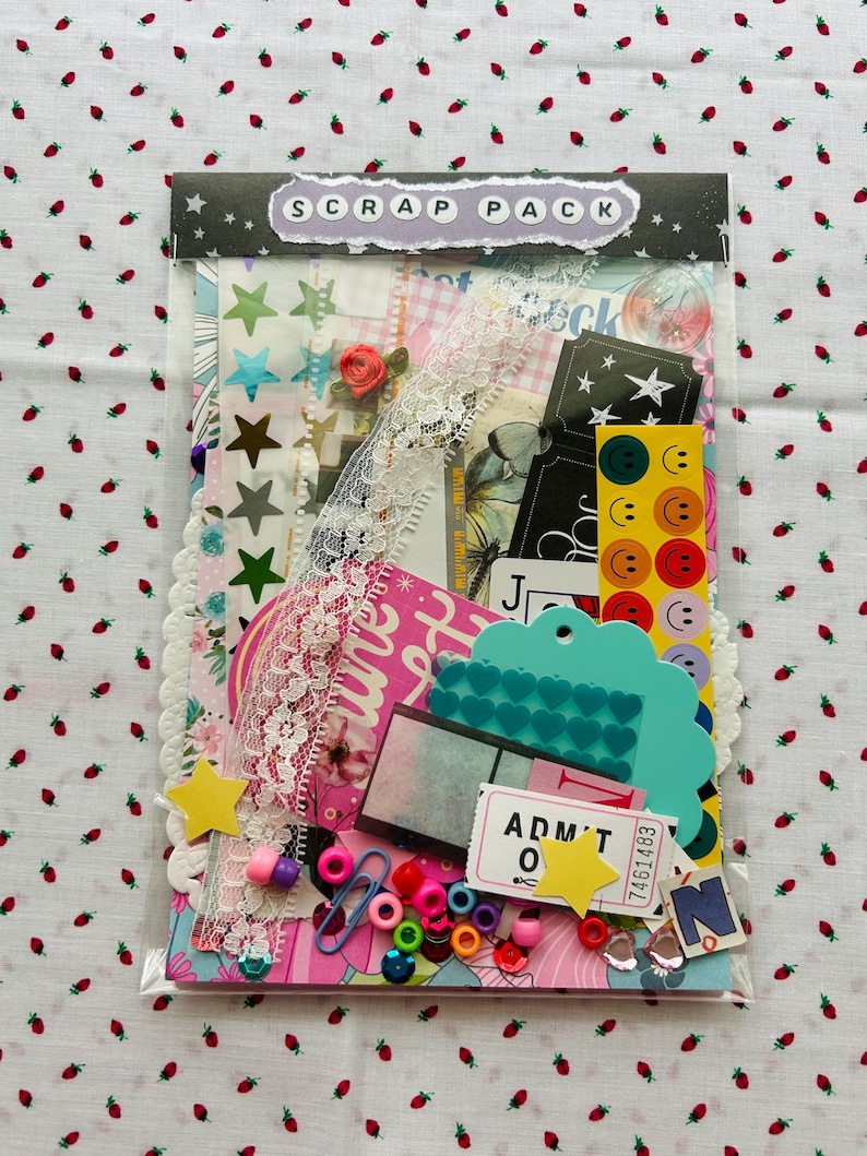 May include: A clear plastic bag filled with scrapbooking supplies. The bag contains various embellishments, including star stickers, lace trim, colorful beads, paper clips, and die-cut shapes. The top of the bag has a label that reads "SCRAP PACK".