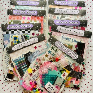 May include: A collection of scrap packs, each sealed in clear plastic. The packs feature a variety of patterned papers, stickers, and embellishments. Each pack is labeled with a purple tag that reads "SCRAP PACK" in white letters. The packs are arranged on a white surface with red strawberry patterns.