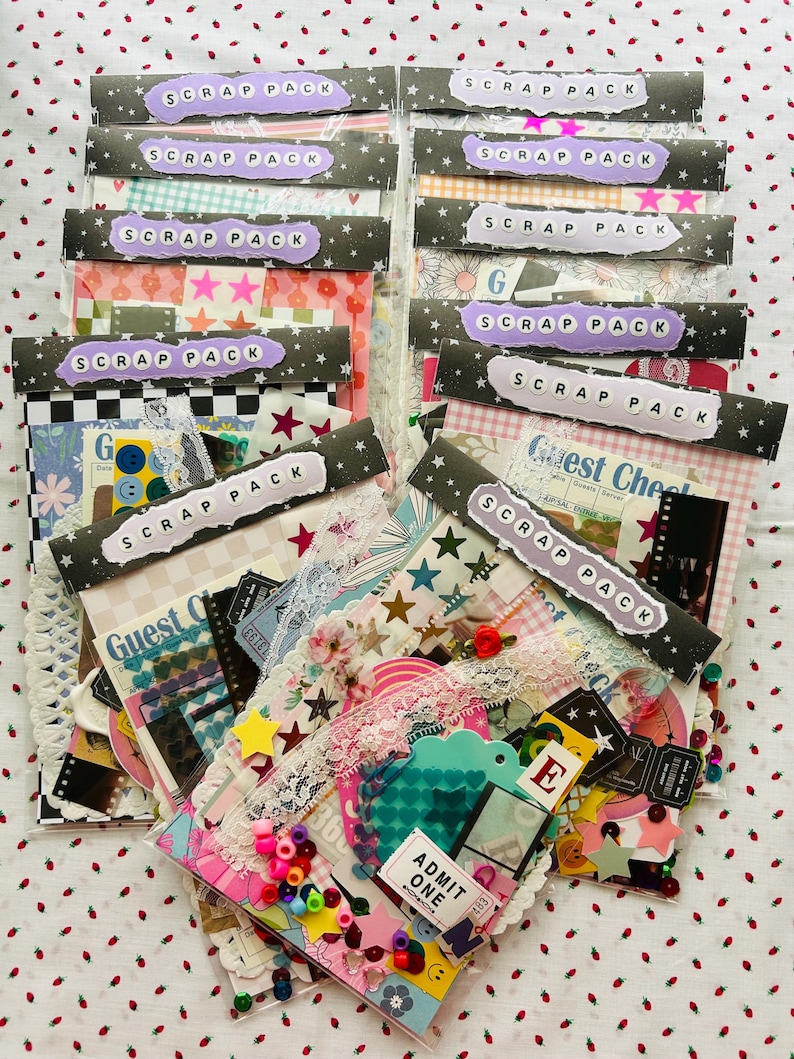 May include: A collection of scrap packs, each sealed in clear plastic. The packs feature a variety of patterned papers, stickers, and embellishments. Each pack is labeled with a purple tag that reads "SCRAP PACK" in white letters. The packs are arranged on a white surface with red strawberry patterns.
