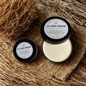 Puede incluir: Dos latas redondas de metal negro que contienen crema de zinc al 40%. La lata más grande está abierta, revelando una crema suave de color blanquecino, mientras que la más pequeña está cerrada. Las etiquetas muestran 40% ZINC CREAM.