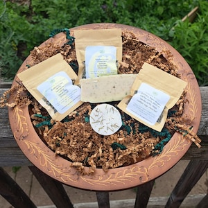 May include: A copper-coloured decorative tray filled with brown paper, green ribbon, and small packages of herbs and soap. The packages have labels with text. The tray has a gold floral design around the rim.