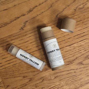 May include: Two tubes of honey tallow lip balm on a wooden surface. One tube is open, revealing the balm, and the other is closed. The labels read "Honey Tallow" and "Local Honey Infused Lip Balm & Multi-Use Face Wax."