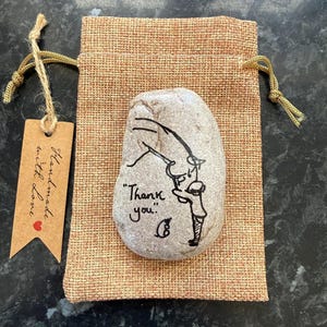 May include: A hand-painted, grey river stone with a black ink drawing of a person holding a gift. The words "Thank you" are written on the stone. The stone is in a small hessian bag with a tag that says "Handmade with love."