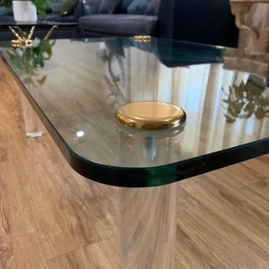 May include: A glass coffee table with a clear acrylic base and gold accents. The table has a rectangular glass top and is supported by four clear acrylic legs.