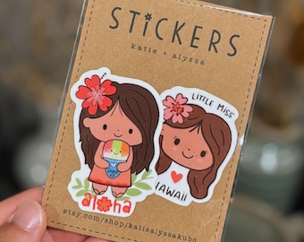 May include: Two stickers on a brown cardstock backing. One sticker features a cartoon girl holding shaved ice with the word "aloha." The other sticker says "Little Miss Hawaii." The cardstock reads "Stickers" and "katie + alyssa."