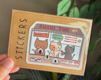 May include: A sticker featuring a sushi restaurant scene with three cartoon cats. The sticker reads "Sushi Time" and "Weekly Special!" The cats are depicted with sushi rolls and nigiri. The sticker is on a brown card that reads "Stickers".