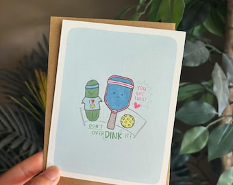 May include: A greeting card with a light blue background featuring cartoon pickle and paddle illustrations. Text includes "Don't Over Dink It!" and "You Got This!" The card is held in a hand, with a brown envelope backing.