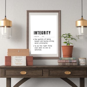 May include: A framed print with the word "INTEGRITY" and its definition, displayed on a dark wood table. The table also holds decorative boxes, books, and a potted plant. Two pendant lights with gold accents hang above.