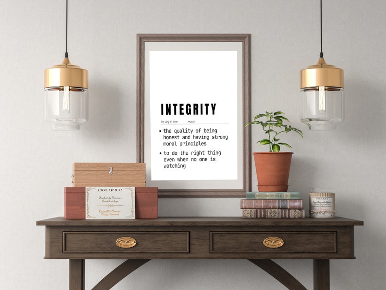 May include: A framed print with the word "INTEGRITY" and its definition, displayed on a dark wood table. The table also holds decorative boxes, books, and a potted plant. Two pendant lights with gold accents hang above.