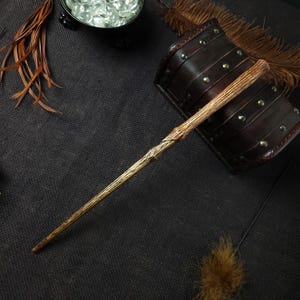 May include: A wooden wand with a textured, naturalistic design. The wand is primarily light brown with darker brown accents, and has a pointed tip. The wand is set against a dark background with other decorative elements.