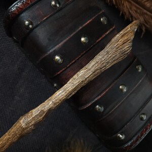 May include: A handcrafted wooden wand with a textured, natural finish. The wand is light brown and appears to be carved from a branch. The background features a dark brown leather bracer with metal studs and a feather.