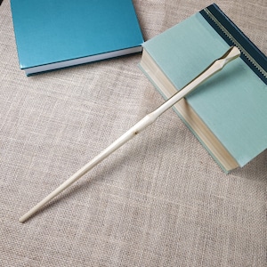 May include: A wooden wand with a light brown colour and a slightly pointed tip. The wand is lying on a beige hessian surface.